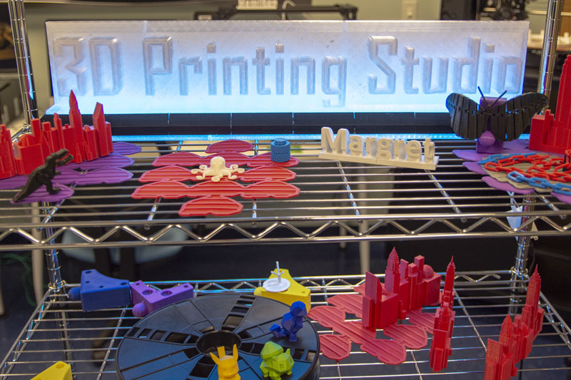 Wire shelf rack with colorful samples of 3D printed objects. A lit 3D Printing Studio sign is behind the models.