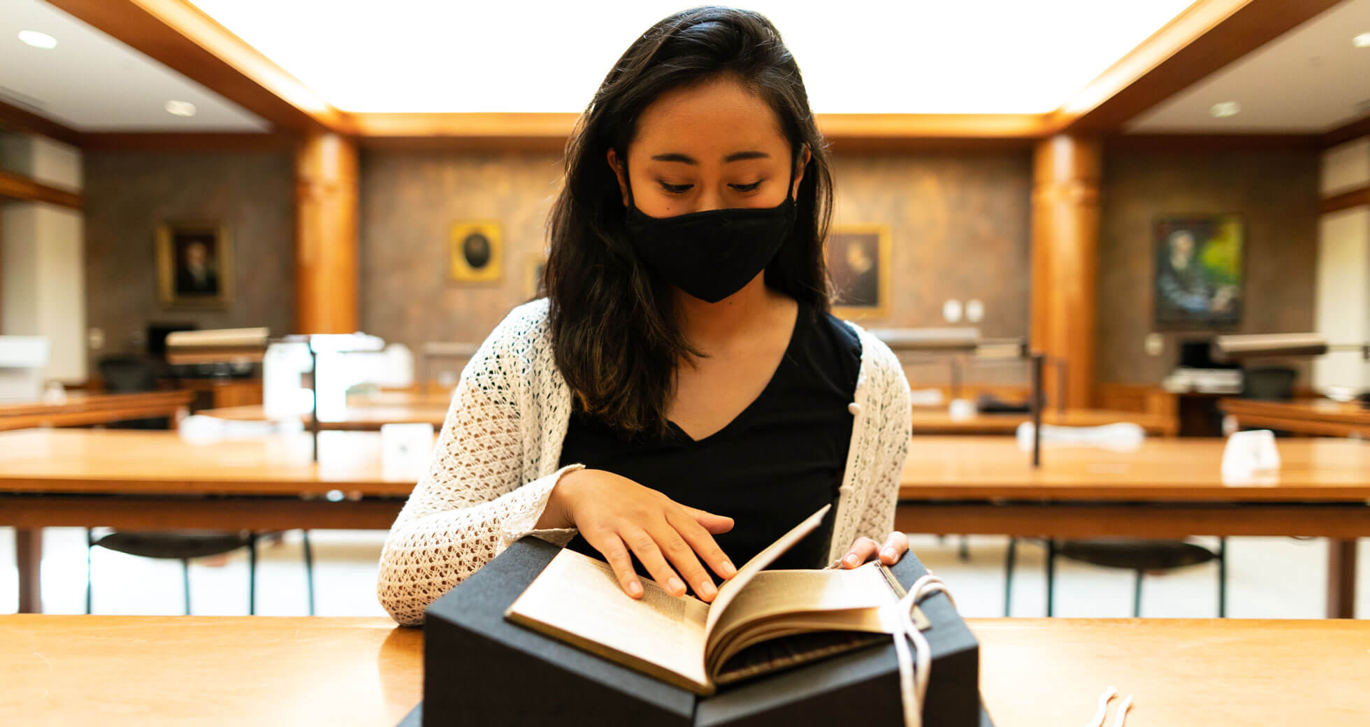 Woman wearing face mask delicately turns the pages of an old book, which is resting on a v-shaped stand