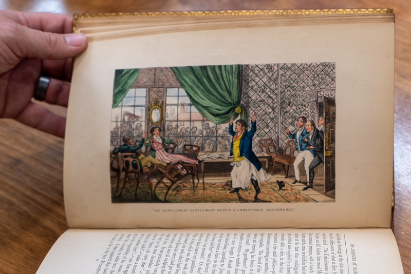 A hand holds a book open to a colorful illustration of a great commotion: a man in a parlor throws up his hands and runs while others fall over a table or run in behind him. A crowd watches through large windows. 