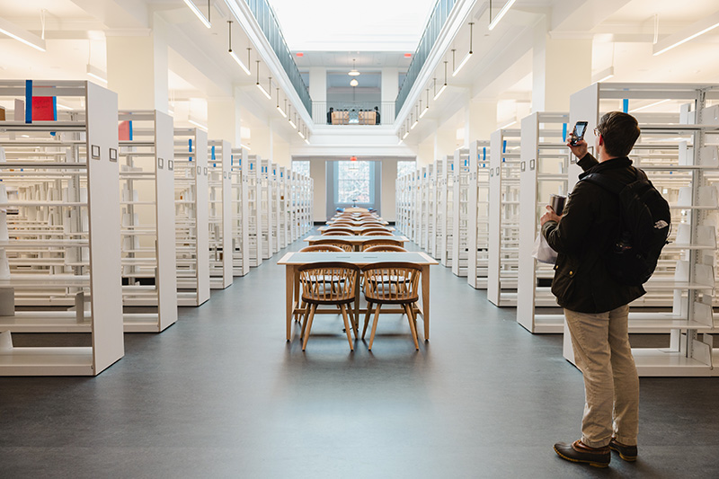 A student taking a photograph in a brightly lit library space with shelving and a row of wooden tables and chairs.