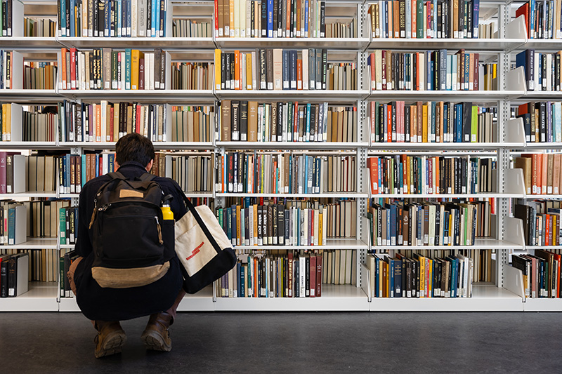 A person wearing a backpack and carrying a tote bag squats, reading, in front of a shelf of books in a library.