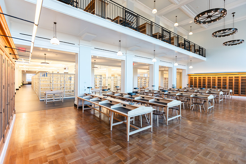 A reading room in a library with parquet-style floors and a double-height ceiling. Shelving can be seen in the background and seating on the floor above behind a railing looks down into the room.