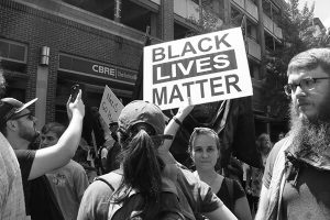A woman in a crowd holds a Black Lives Matter sign over her head. She is looking directly at the camera. People around her are looking in different directions, watchful. The CBRE Charlottesville sign is visible in the background.