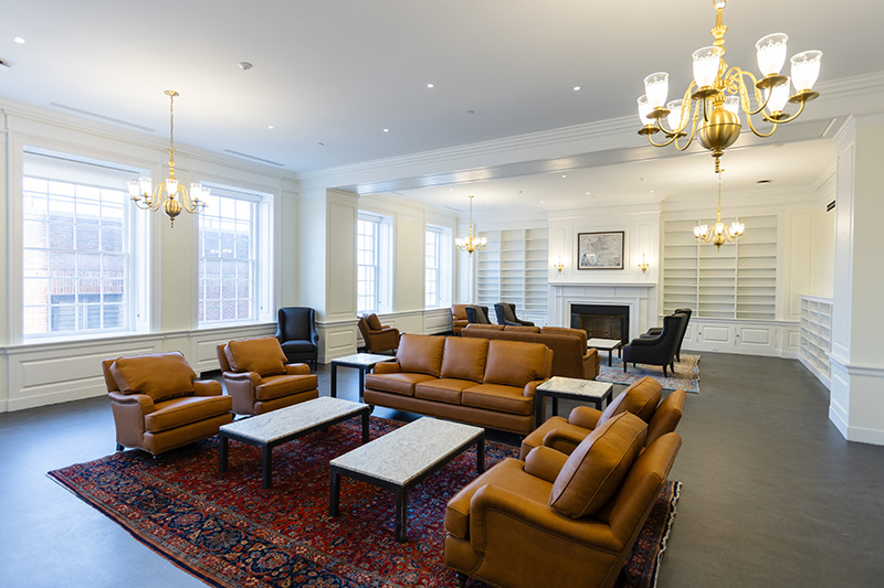 2 area rugs with tables and soft seating on each rug create 2 separate sitting areas in a large room. The room has a fireplace, large windows, chandeliers, and shelving on either side of the fireplace.