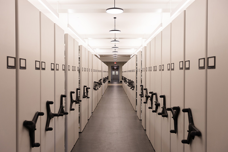 View down an aisle between two rows of compact or mobile shelving, with handles on the end of each range of shelving.