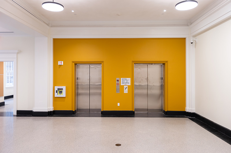 A wall painted a honey mustard yellow with two stainless steel elevator doors side by side.