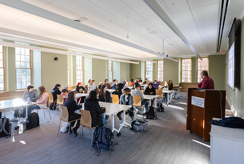 A large bright classroom full of students sitting at tables while a person stands at a lectern in front of a screen. On the lectern is a “UVA Library” logo.