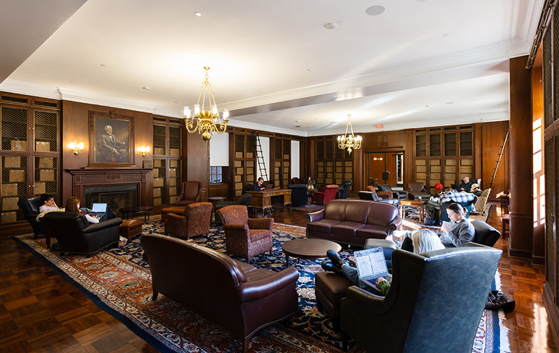 A large but cozy-looking wood paneled room with people sitting in soft seating. The room also has bookshelves, tables, and a fireplace.
