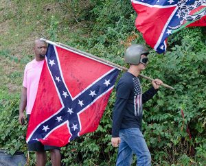 A person wearing a motorcycle helmet and goggles carries a Confederate flag past a man who is looking at him with a direct stare and serious brow.