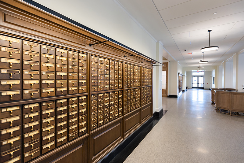 A row of card catalog drawers line the left side of a hallway, which is broken by an opening. At the end of the hallway are two doors, and the space is lighted by fixtures hanging from the ceiling down its length.