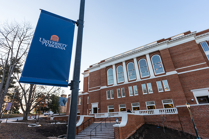 A blue flag with a University of Virginia logo attached to a pole is in the foreground of a photo showing a multiple-story brick building with white pilasters between large arched windows, many smaller windows, and stairs leading up to terrace space.
