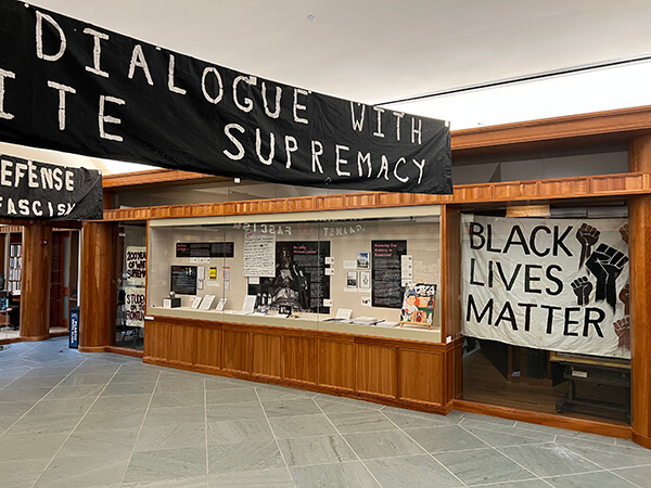 A view of the exhibit space shows a display case, large Black Lives Matter sign, and portions of large black overhead banners showing words like ‘facism’ and ‘white supremacy.’