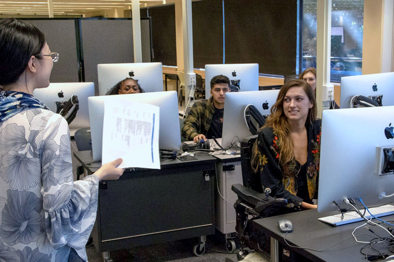 An instructor is talking to a group of students as they each sit in front of a desktop computer.