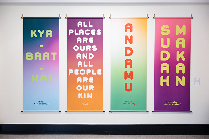Four colorful banners hang against a white white. They read: Kya baat hai, Hindi, how amazing!; All places are ours and all people are our kin, Tamil; Andamu, Telugu, inner beauty; Sudah makan, Malaysian, have you eaten?