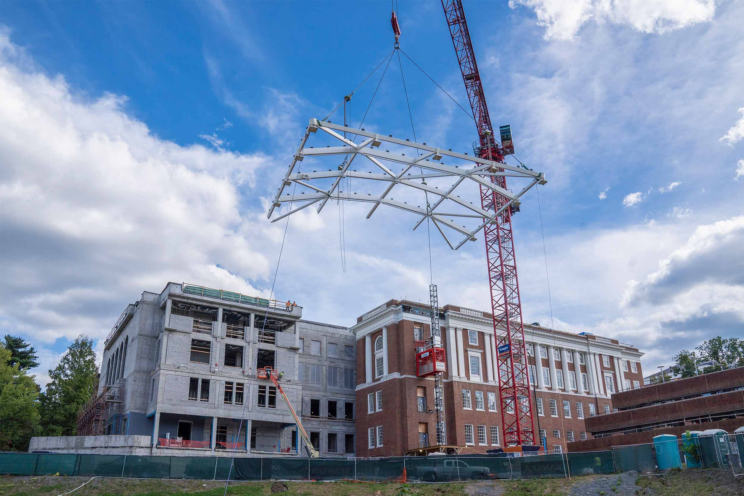 A massive crane lifts a metal structure up and over a rectangular building which is partially under construction