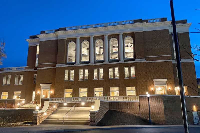 A nighttime photo of multistory brick building with large windows illuminated from within.