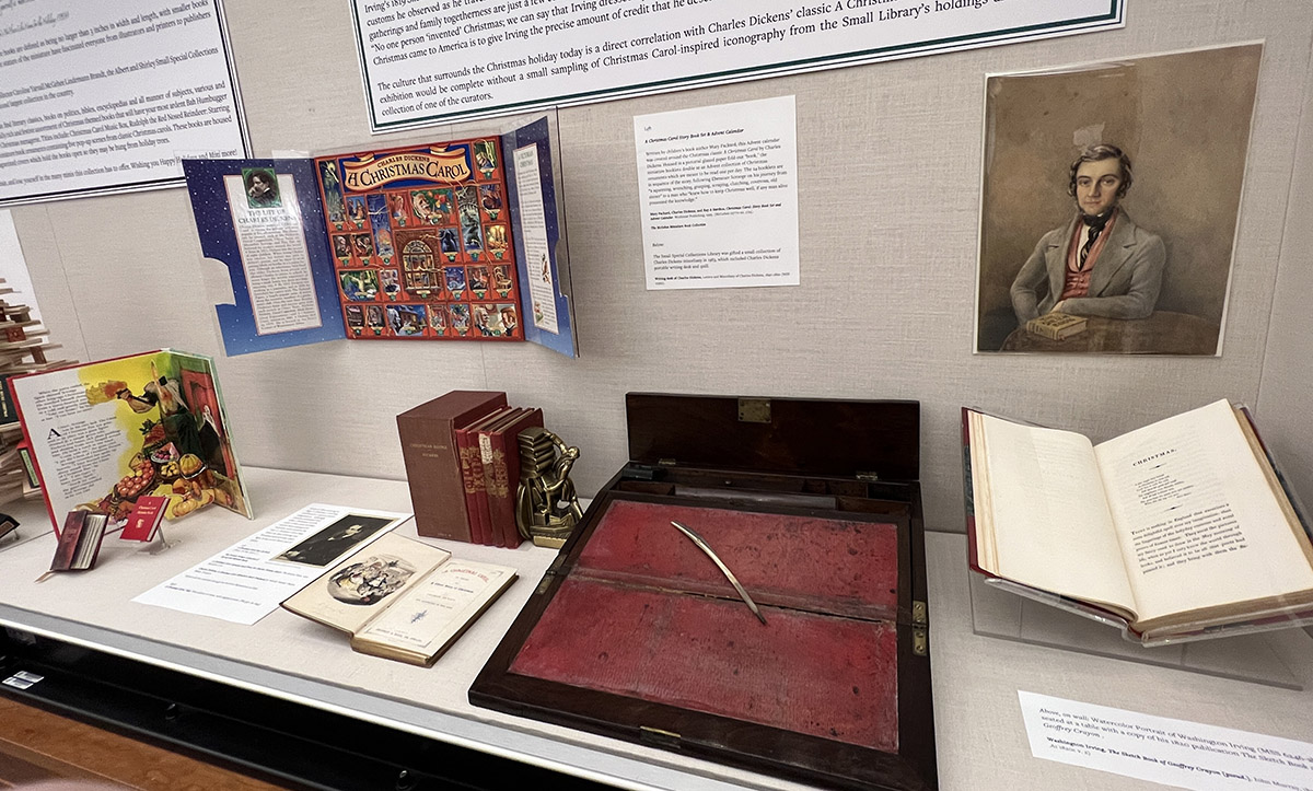 An open glass exhibition case with an advent calendar, books, a portrait, and a small writing table inside. 