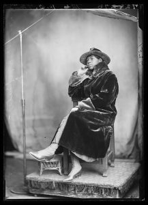 Black and white photo of a Black woman posing in a chair wearing a luxurious velvet coat with fur lining, lace-up boots, and a flat-brimmed hat