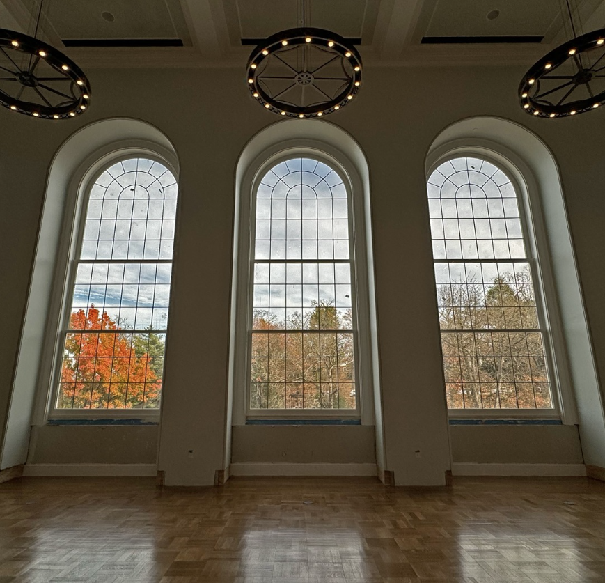 Wagon wheel lights hover over tall windows overlooking a wooded area. The room has wood floors