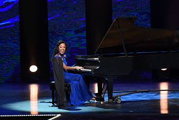 A woman in an elegant blue dress plays a grand piano on a dramatically-lit stage, as she looks directly at the audience and smiles