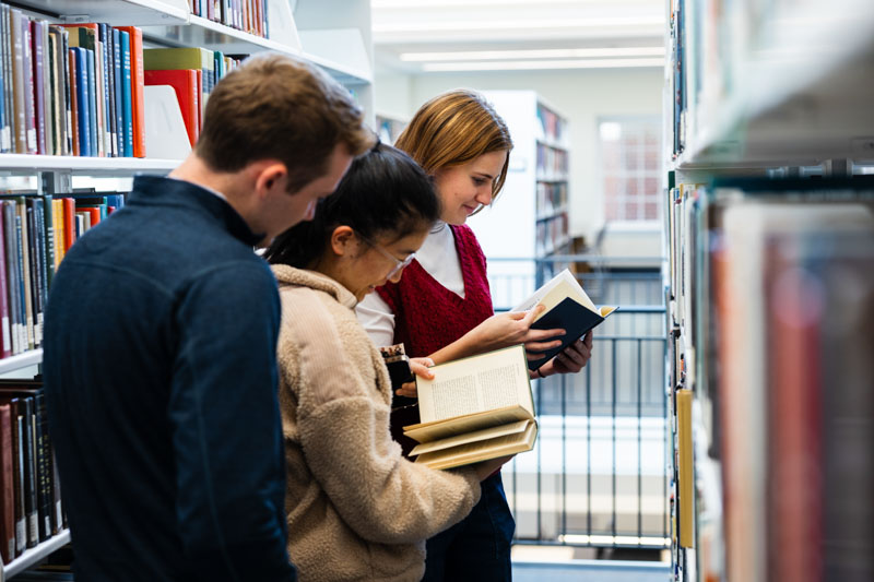 Three smiling students look at books while standing in the stacks.