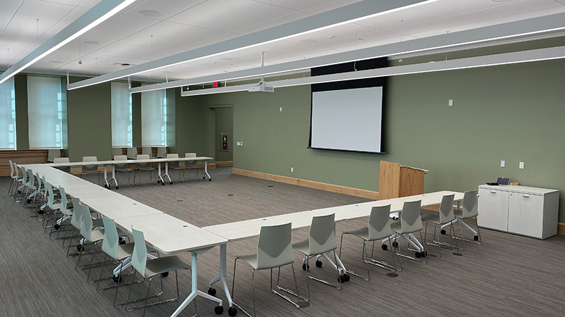 A large room with tables in a U shape around an open center space. A screen is on the wall for projecting.